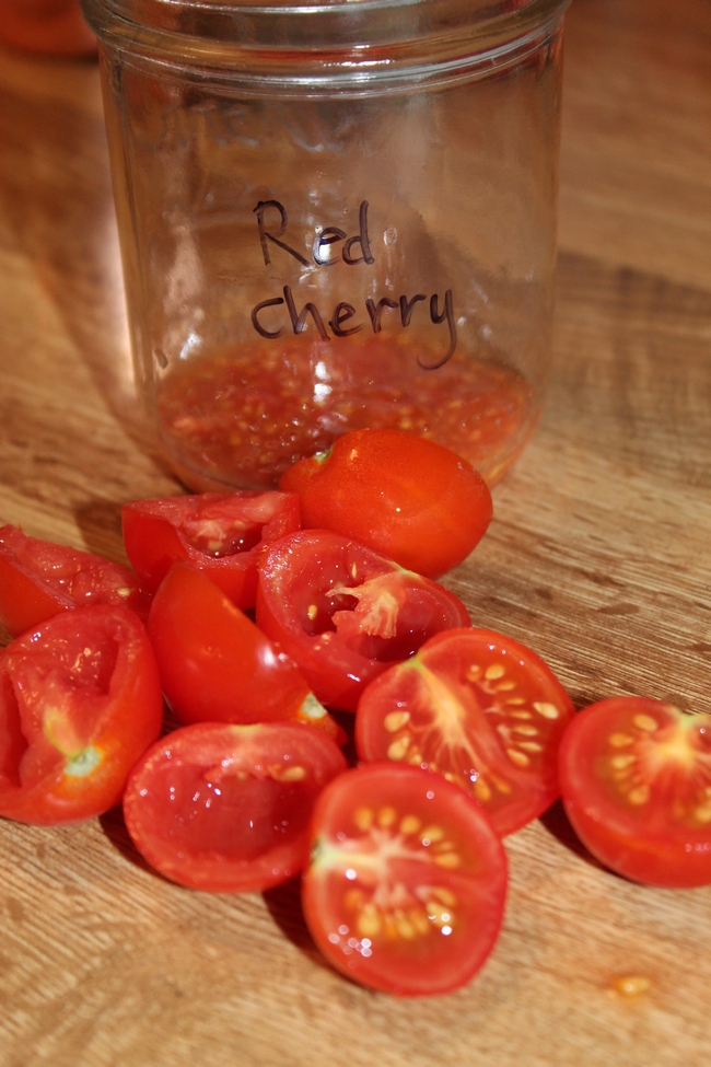Sliced cherry tomatoes next to a glass container with cherry tomato seeds.