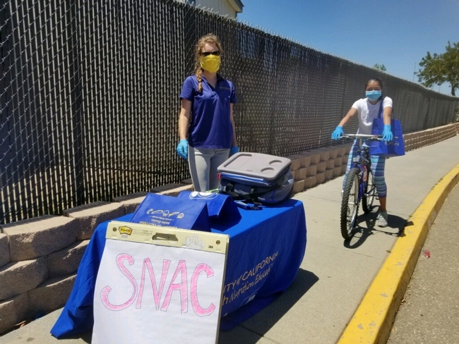 Table set-up on a sidewalk in front of a tall fence. The table has a blue table cloth with a University of California logo. Woman with a blue shirt, latex gloves, and yellow face covering is looking at the camera. Behind her is a girl on a bicycle with a blue bag of materials over her shoulder.