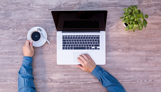 An overhead shot of a person with one hand holding a coffee mug and the other hand on the mousepad of a laptop.