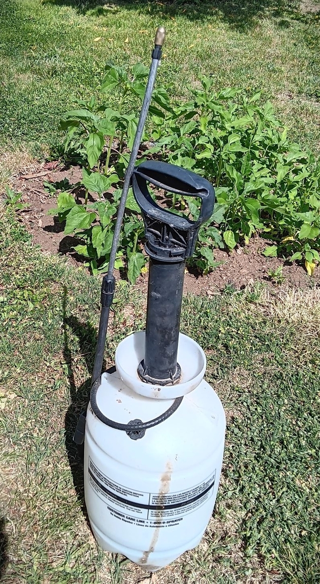 A hand-pump sprayer with the handle extended and spray nozzle resting against the tank, surrounded by lawn.