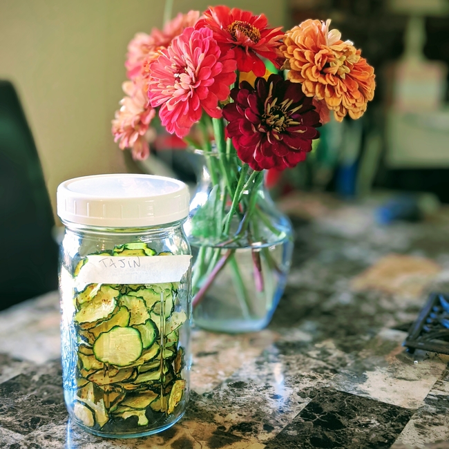 A jar containing zucchini chips next to a vase of flowers.