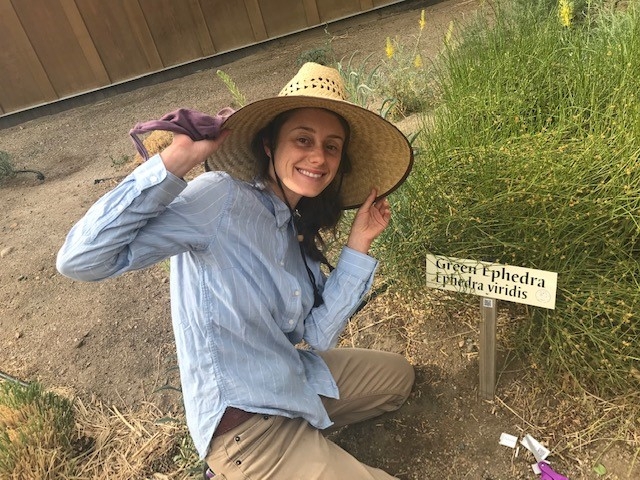 Woman in hat in front of plant with sign.