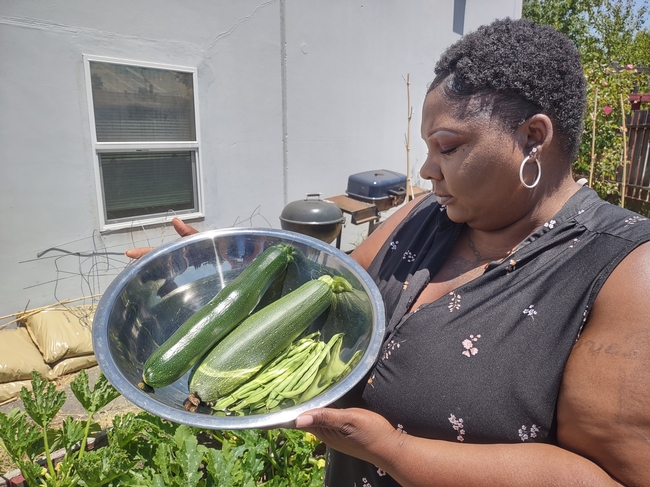 A Black woman, standing in a garden, holds a stainless steel bowl filled with zucchini and beans