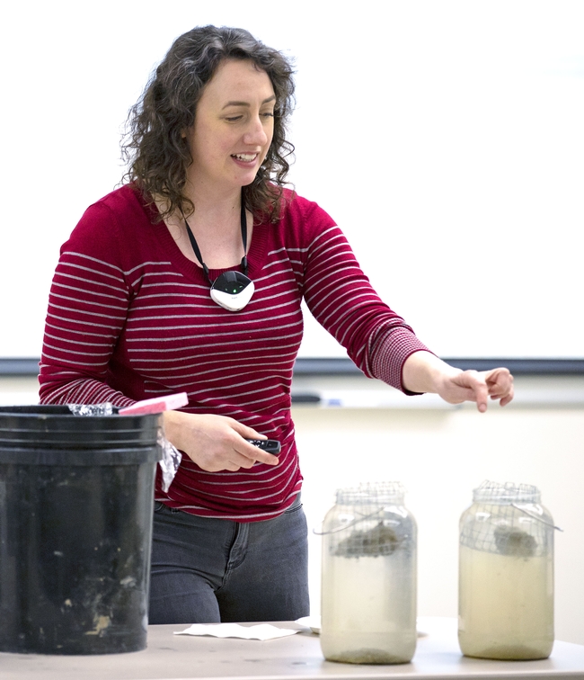 A woman points to a jar filled with water that is less cloudy because the clump of soil in it is holding together
