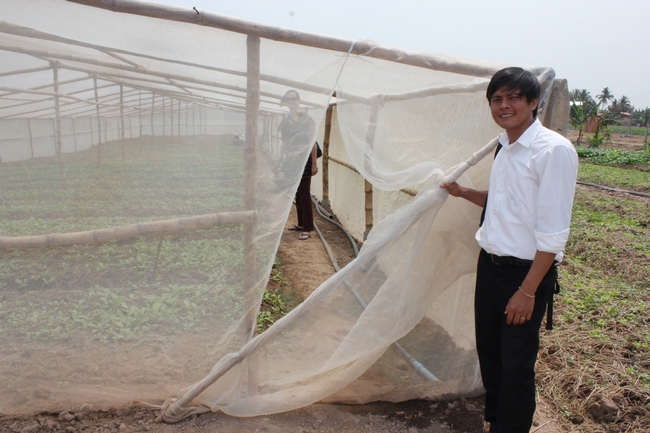 Outside, a young man holds open a net door into a simple bamboo structure covered in thin netting. Inside the nets, a woman stands among the vegetable seedlings in the ground.