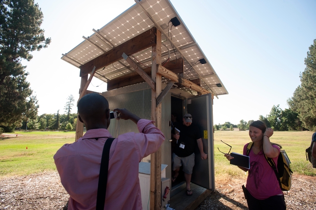 People with notebooks in hand explore a small shed with solar panels on the roof, while one man takes photos of the technology.
