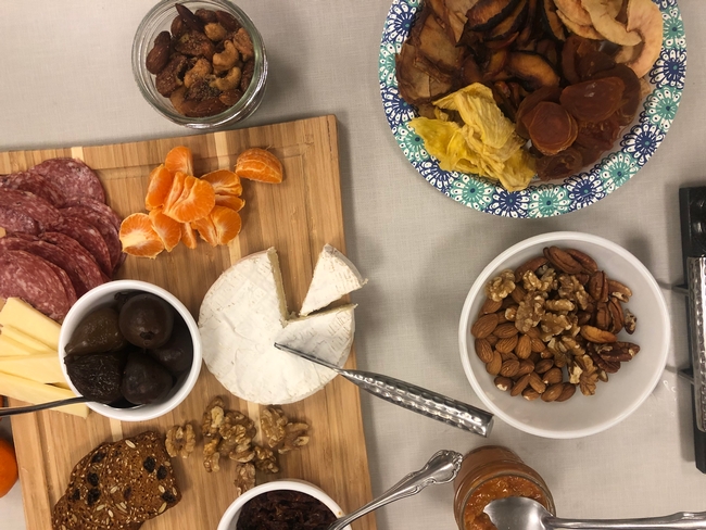 Mixed nuts in bowls shown with a wheel of brie, mandarin segments, salami and dried fruit.
