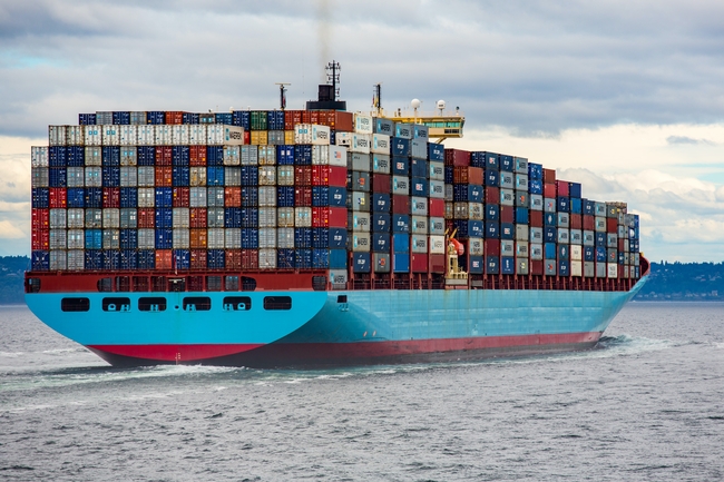 A ship piled high with shipping containers sails out of a port.