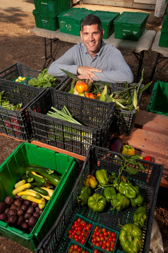 Ned Spang, surrounded by plastic bins of fresh produce, poses smiling with his arms crossed on the edge of a bin containing apples and corn on the cob.