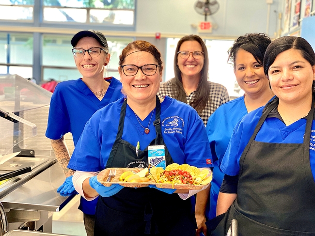 Vanessa Zajfen, wearing a hairnet and surrounded by four colleagues, holds a school lunch tray displaying a meal at based on fresh products. width=