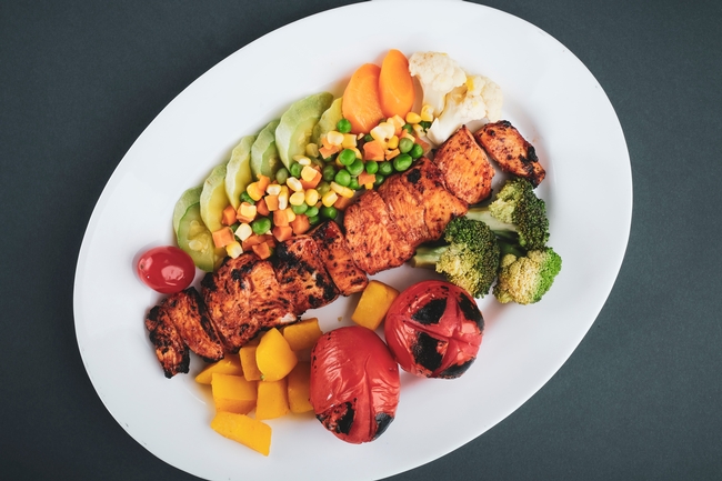 Grilled boneless chicken surrounded by broccoli, squash, corn, peas, carrots cauliflower and tomatoes on a white plate.