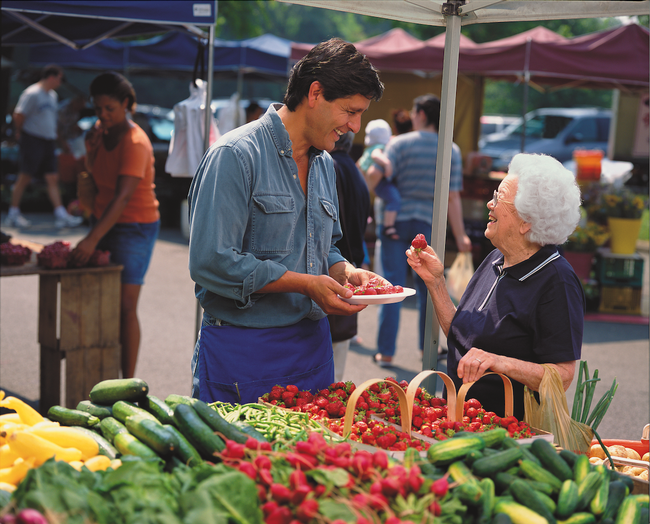 A white-haired woman holds a strawberry and smiles at a brown-haired man holding a plate of strawberries at a farmers market .