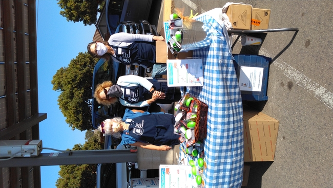 Three people with blue aprons in a parking lot standing in front of a table with information and materials