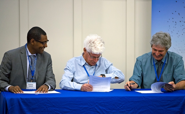 Three men sit at a table signing documents