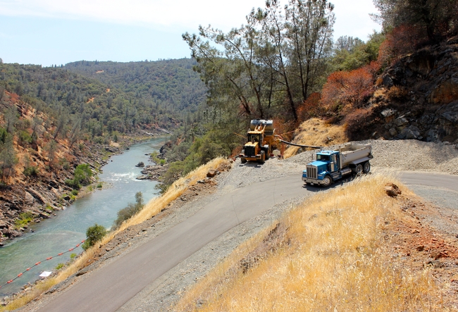 Trucks haul gravel from a nearby quarry, heavy equipment is used to load the material, pipes deposit the aggregate into the river.