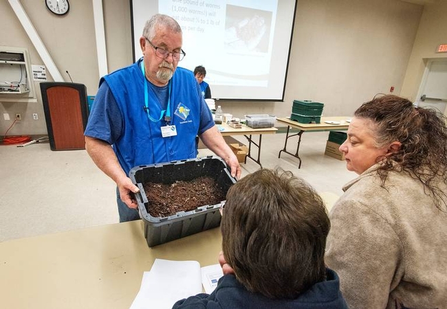 Ted Hawkins wearing a blue shirt, blue MG vest and MG lanyard around his neck while holding a black bin with worms and dirt while students look on.