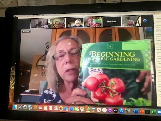 A Zoom meeting screenshot of Nancy Herz holding up a Beginning Vegetable Garden book with a hand holding a vine of red tomatoes.