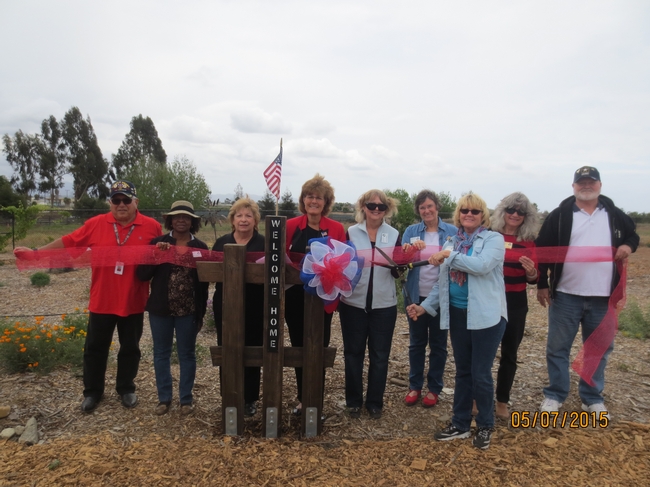 Nine people stand on a wood chip covered garden path, behind a post, engraved vertically with the words welcome home.” A small American flag is mounted atop the post. One person holds garden shears, poised to cut through a translucent red ribbon being held by eight other gardeners.