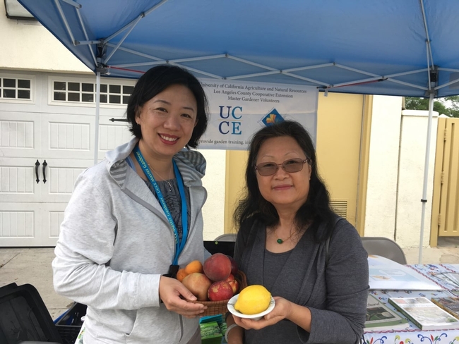 Jennifer Kwoon stands with a community member, both holding stone fruit and citrus, in front of a table at an outdoor farmer's market.