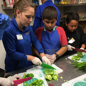 An adult with gloves on cutting a green bell pepper next to a youth with an apron watching the process