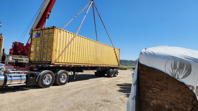 A crane loads a yellow shipping  container filled with farm equipment onto a flatbed trailer