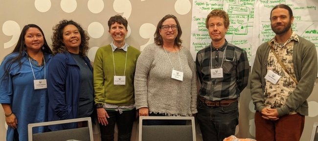 Six people wearing name tags stand in front of a wall that is partly covered in white poster paper with discussion notes written in green ink.