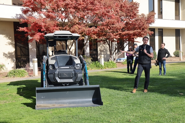 Standing on a lawn, a man gestures toward a piece of machinery.