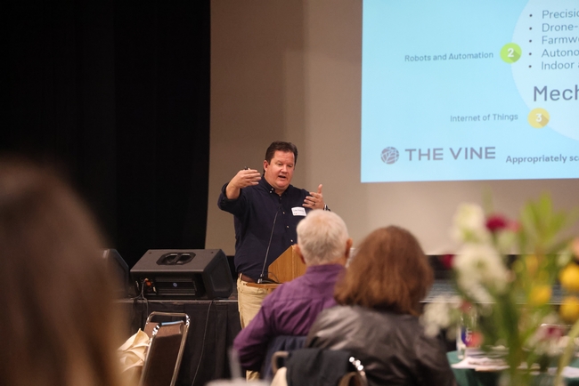 Youtsey speaks to a group in a conference room. A PowerPoint slide projects points about The VINE.