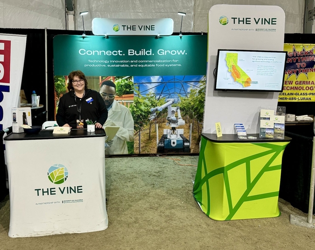 Terri stands behind a table with The VINE logo. Sign behind her reads: Connect. Build. Grow.