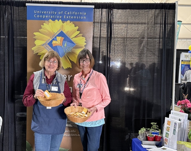 Two volunteers hold baskets of seed packets while standing in front of a Master Gardener banner.