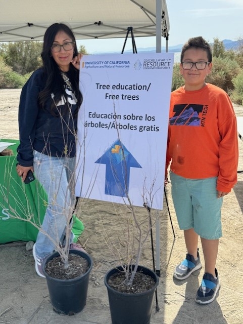 A woman and boy stand beside a sapling and sign that reads: Tree education/free trees. Educacion sobre los arboles/arboles gratis.