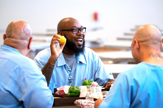 Three bald men wearing blue prison shirts sit at a table eating lunch.