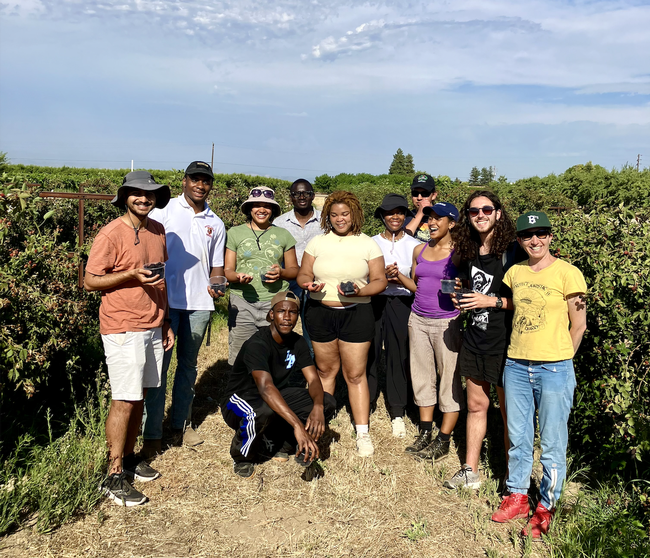 Eleven young people holding clear plastic cups of blackberries pose between rows of blackberry bushes.