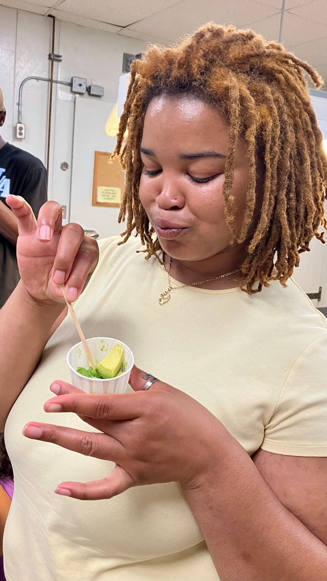 Using a wooden toothpick, a young Black woman spears a morsel of green avocado in a paper cup.