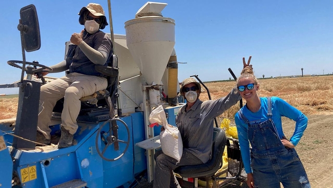 A person sits in the driver's seat of a combine, another person sits beside the combine holding a bag in his right hand while making bunny ears with his left hand behind Maya's head as she stands beside him.