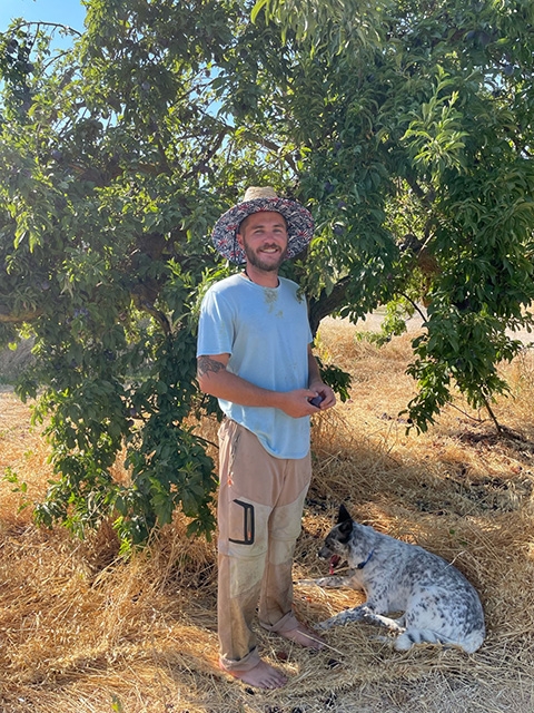 A man stands next to a tree as his dog lays at his feet.