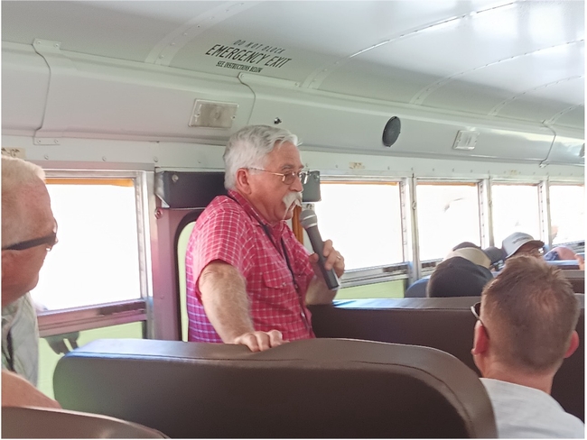 Man in red checked shirt speaks into a mic on a bus.