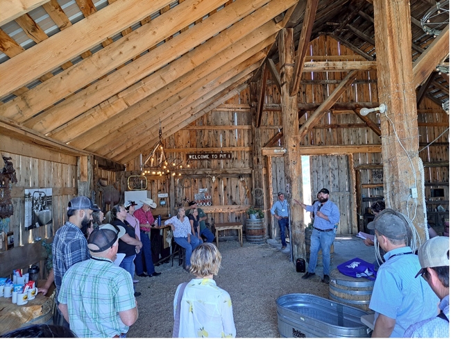 People stand around a man speaking in an open beam barn.