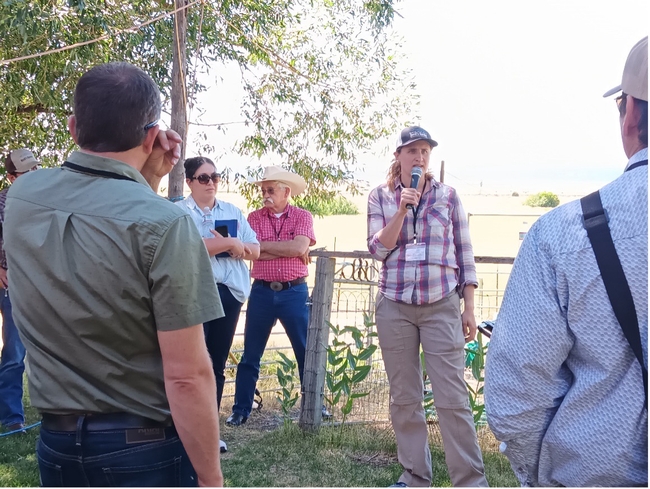 Woman standing in a garden speaks into a mic while people stand around her.