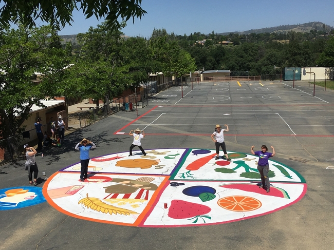 5 educators flex their biceps while standing in a huge colorful MyPlate stencil in a playground.
