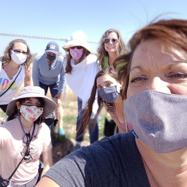 Seven people wearing facemasks and sportswear gather for a selfie outdoors.
