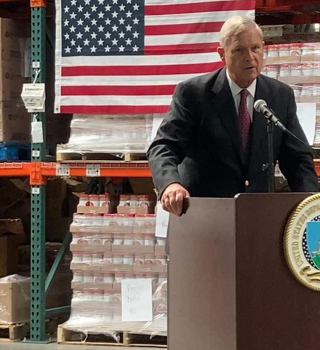 Vilsack stands at lectern with pallets of canned goods on shelves behind him.