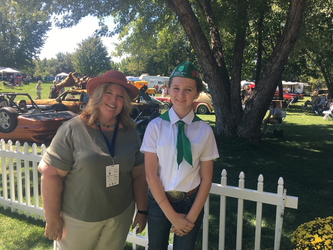June, wearing a hat, stands under a leafy tree beside Lily, who is wearing a 4-H uniform of green and white. Classic cars are in the background.