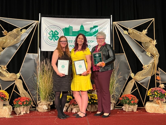Three women pose with plaques in front of decorative autumn backdrop and 4-H conference banner.