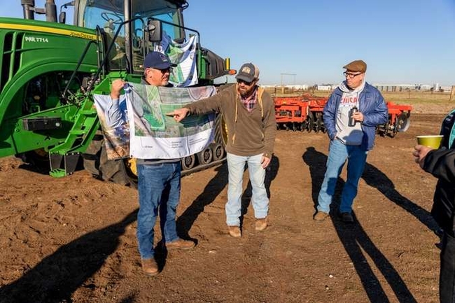 Three men stand on tilled soil looking at a diagram of the field. Green and orange tractors are parked in the background.