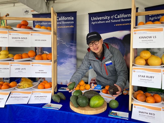 Wearing a UC ANR hat and standing at the ANR booth, Mike holds different varieties of avocados in each hand.