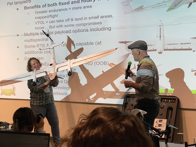 Becca holds a large model airplane as a man wearing a hat speaks into a handheld microphone.