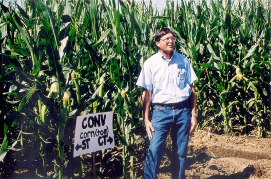Kent Brittan stands at the edge of a field of corn that is taller than he is.