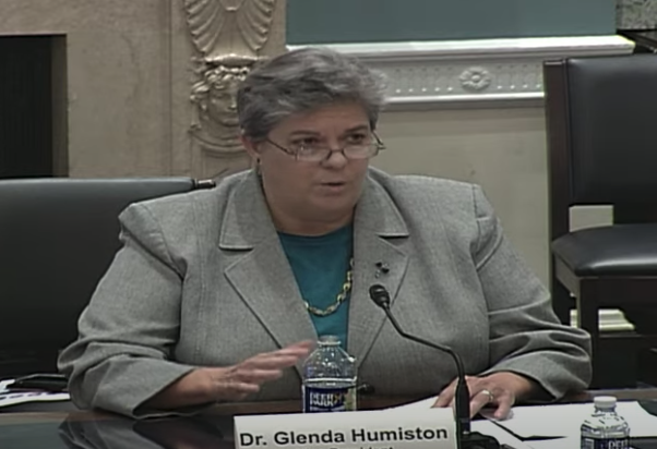 Close up of Humiston sitting at a table speaking, a bottle of water sits near her right hand. She is wearing a gray suit jacket over a turquoise top.