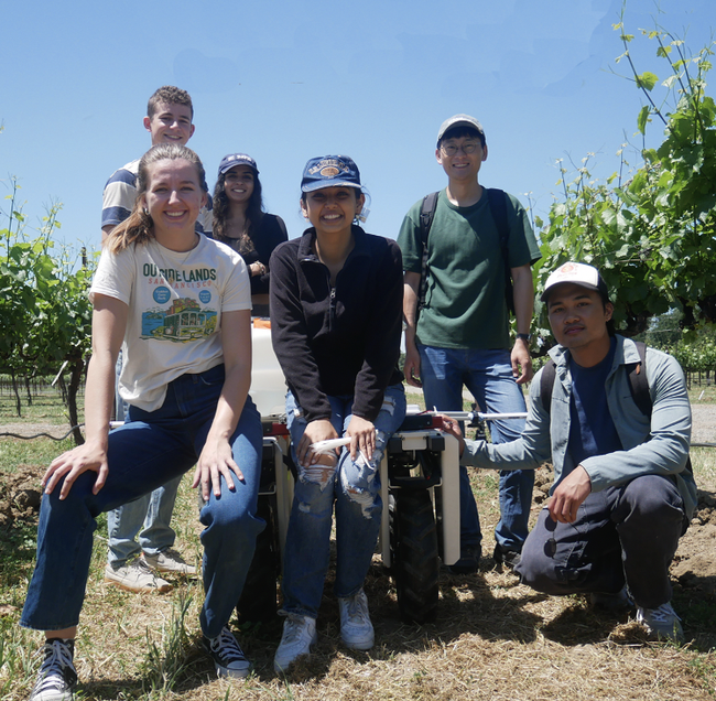 Six people huddle around their robot in a vineyard.